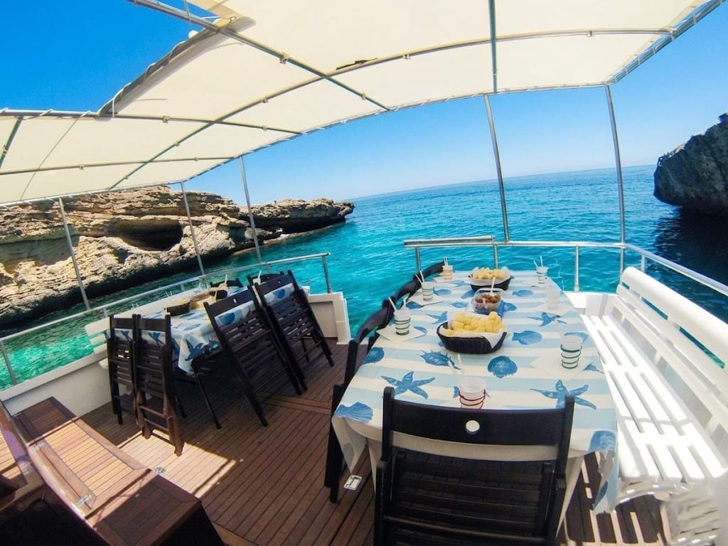 Tavola apparecchiata con tovaglie a tema marino e cestini di pane sul ponte di una barca per escursioni ad Alghero, ormeggiata in una caletta con acque turchesi e grotte marine sullo sfondo.