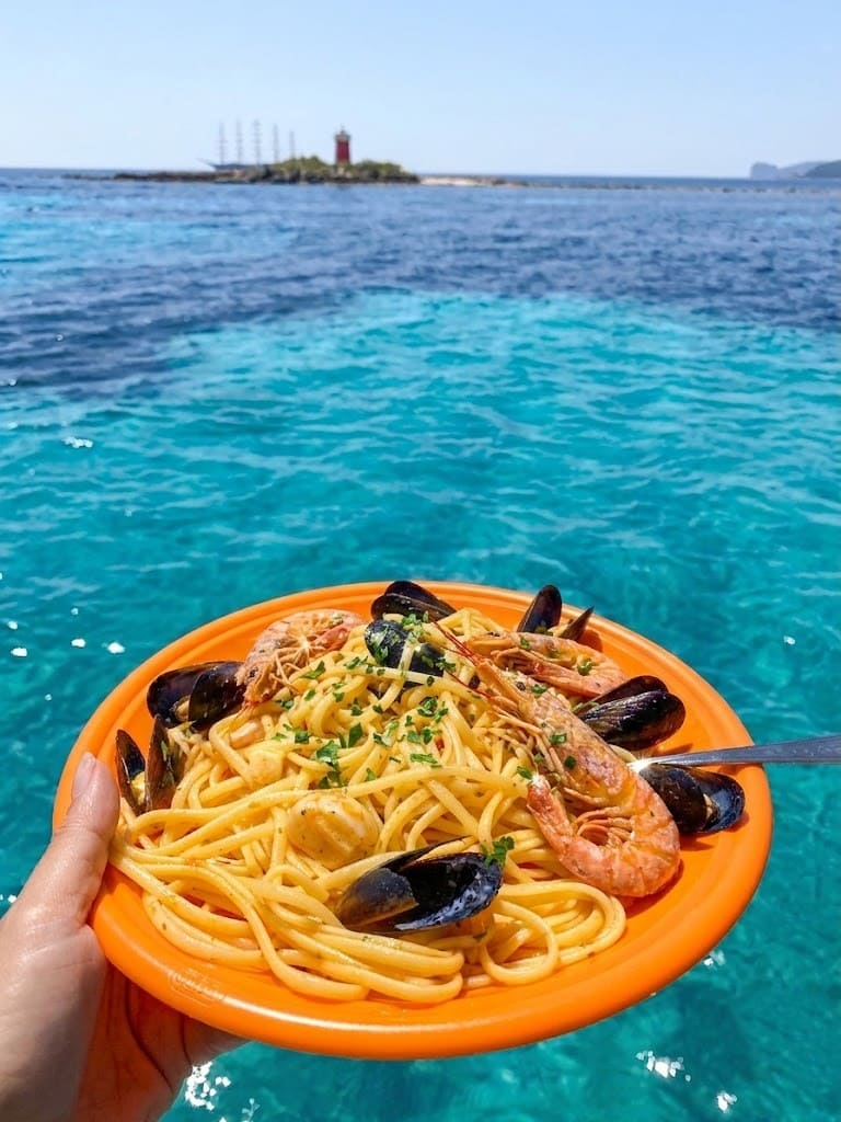 Primo piatto di pasta con gamberoni e cozze fresche servito a bordo durante un'escursione in barca ad Alghero, con il mare cristallino e il faro sullo sfondo.