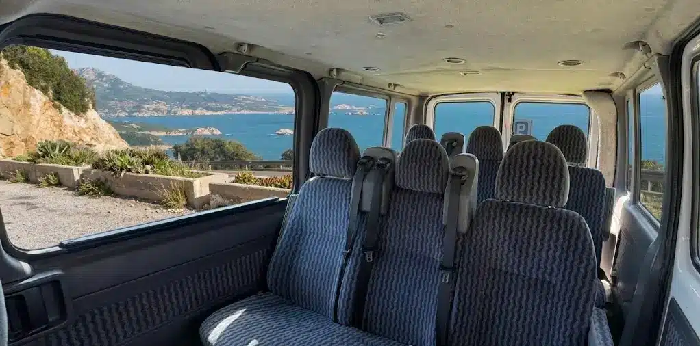 Interno di un van passeggeri con sedili blu e grandi finestrini che incorniciano una vista panoramica sulla costa rocciosa e sul mare turchese di Alghero.